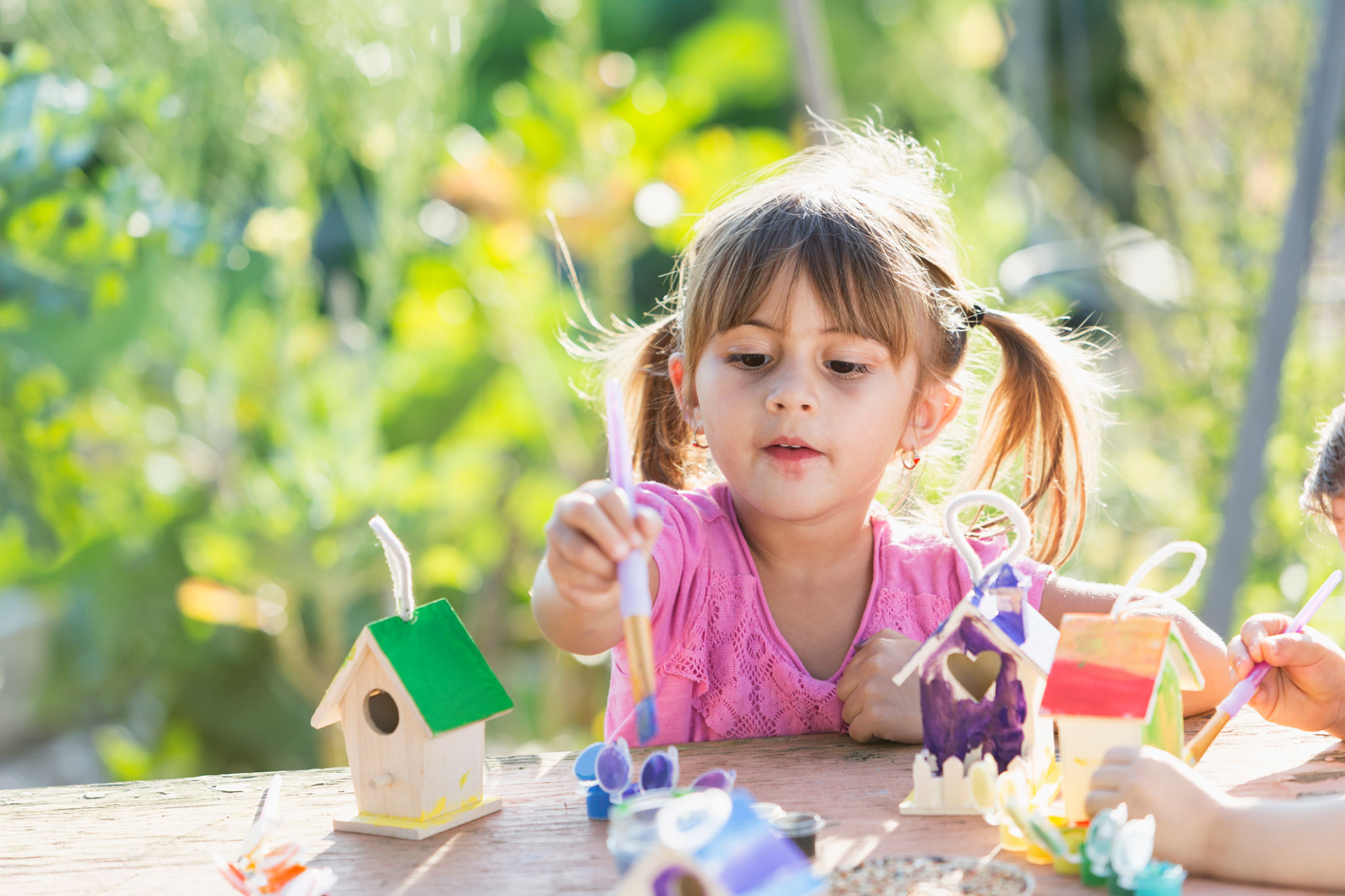 A cute little Hispanic, 3 year old girl sitting outdoors at a table painting a wooden bird feeder.  It is a bright, sunny day in a garden, plants out of focus in the background.  The child is wearing a pink shirt and her hair is tied up in pig tails.